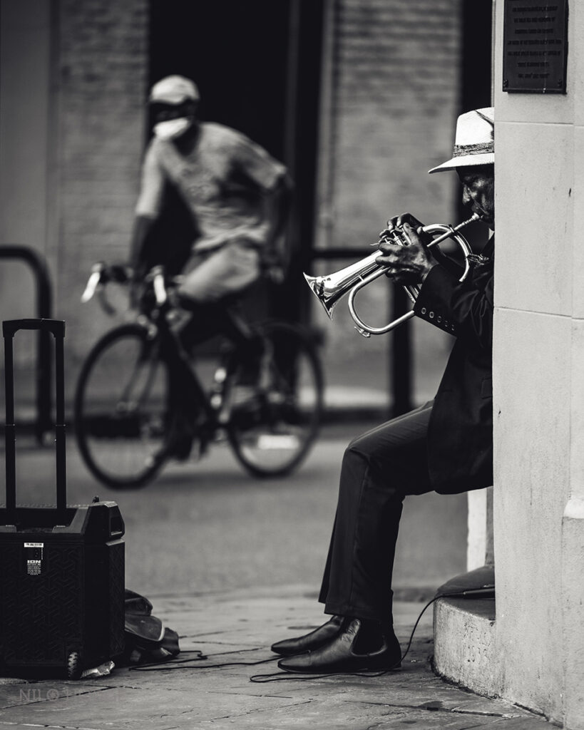 Man sitting on stoop playing the trumpet wearing a fedora as a man wearing a mask rides his bike past in the background